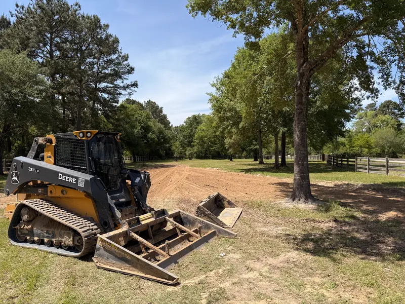 Compact track loader fitted with a land plane sitting beside a freshly cut drainage swale on a wooded residential lot, board fence and oaks behind it
