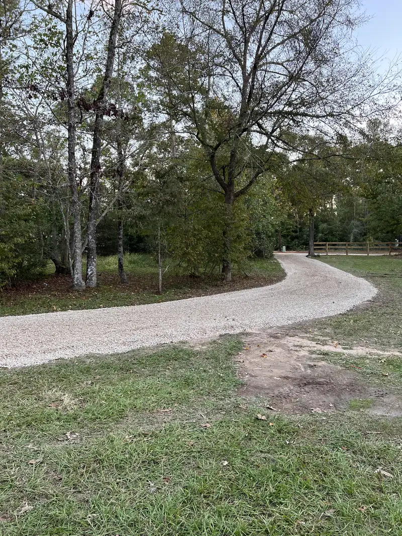 Pale crushed limestone driveway curving away through mature hardwood trees toward a split rail fence in the evening light