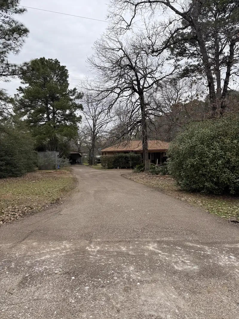 Finished milled asphalt driveway running up to a low ranch house between hedges and bare winter trees