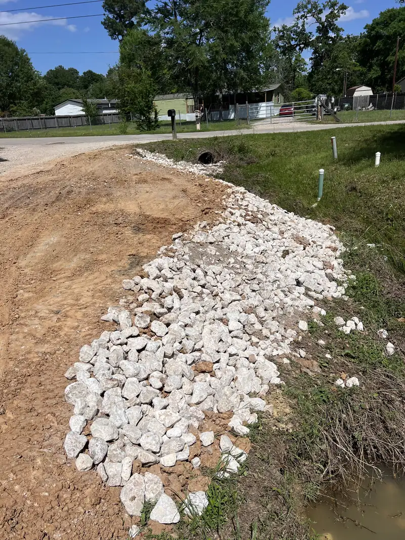 Bank of white limestone rip rap armoring a roadside ditch beside a new dirt drive, with a culvert outfall and standing water at the bottom