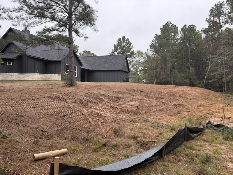 Freshly imported fill dirt graded flat across the slope behind a dark two story house, silt fence running along the low edge