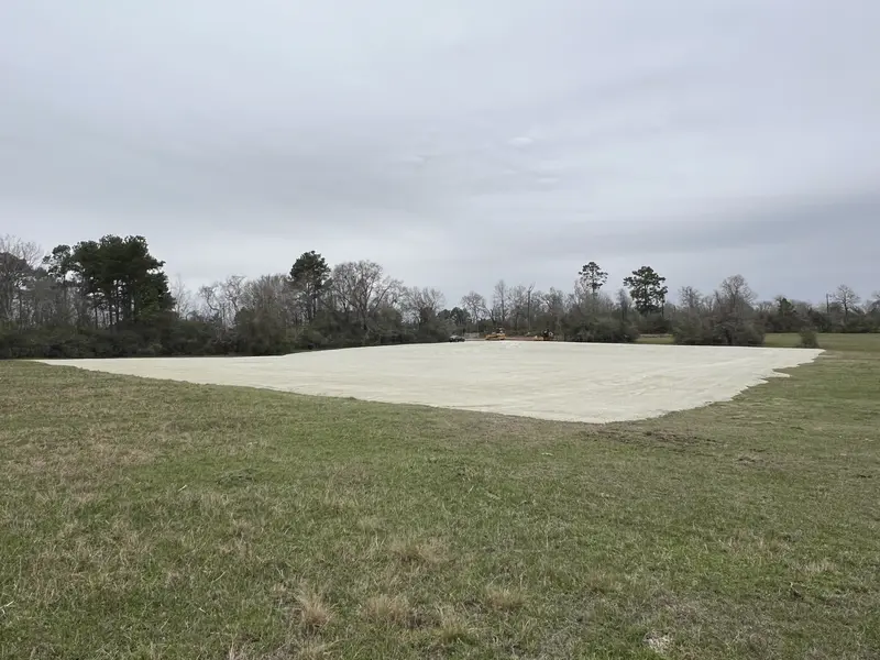 Wide pale compacted lay-down yard built out into a green pasture with a treeline behind it under an overcast sky