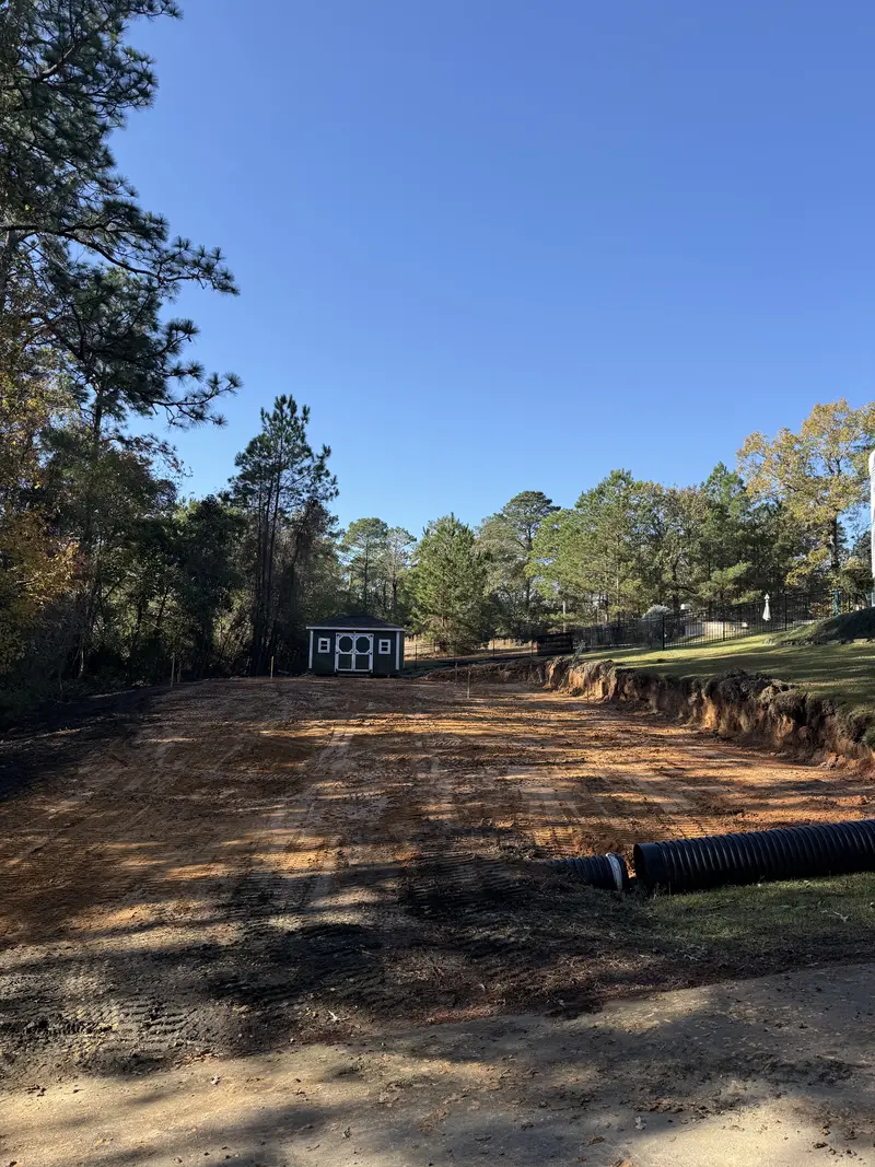 Finished building pad graded level with a black corrugated drain pipe laid along the cut bank and a small shed at the treeline