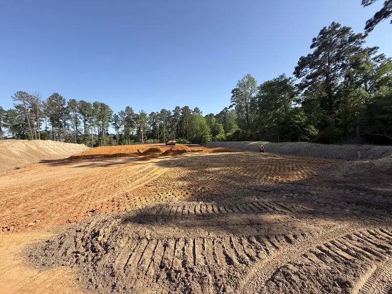 Large orange clay building pad cut between banks with deep tire tracks across it and a dozer working at the far end