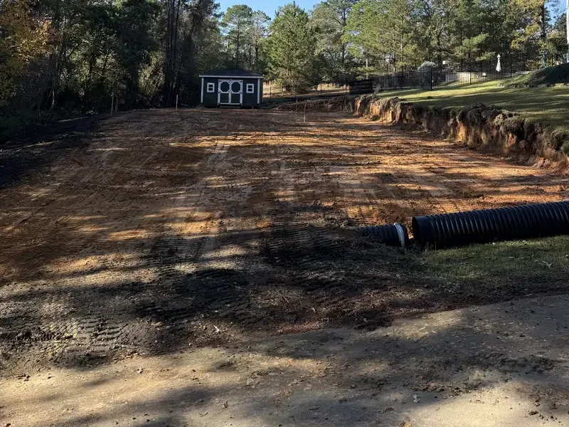 Freshly graded building pad in fall light with a black corrugated drain pipe laid along the cut bank and a small green shed at the treeline